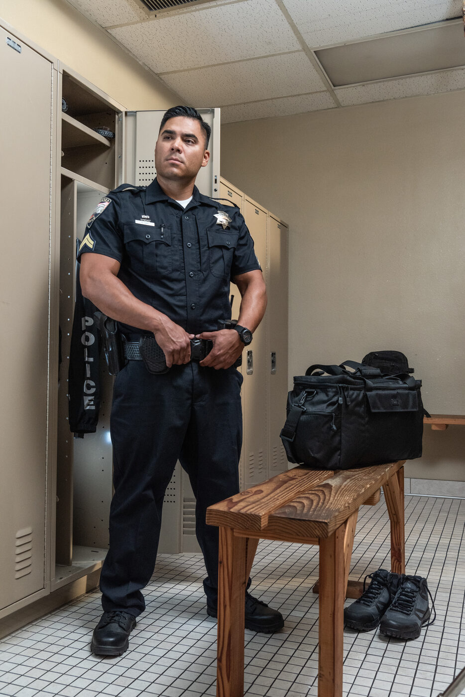Officer in uniform standing in a fitting room with duty boots and a gear bag on the bench